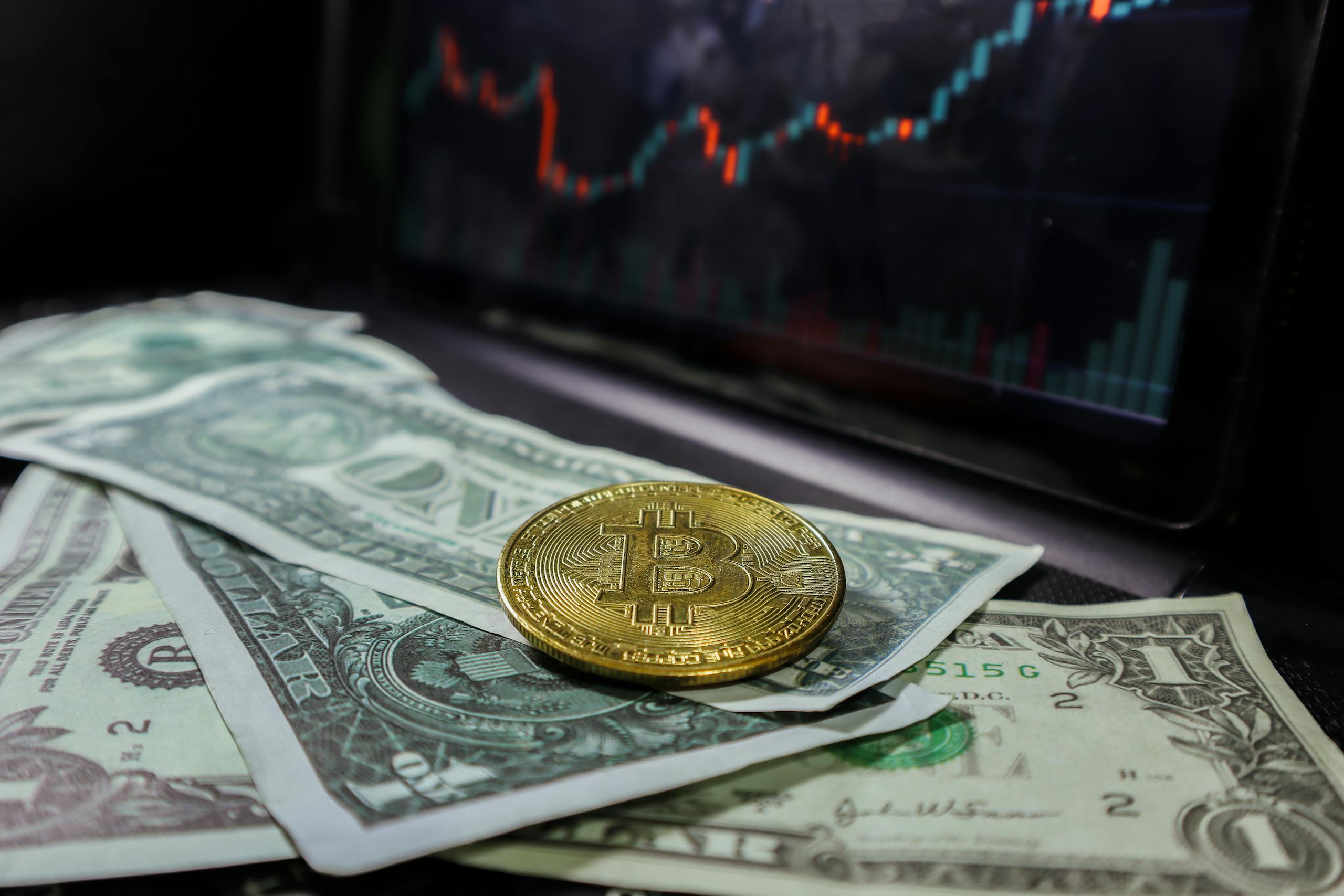 A bitcoin coin placed on US dollar bills with a stock market chart in the background.